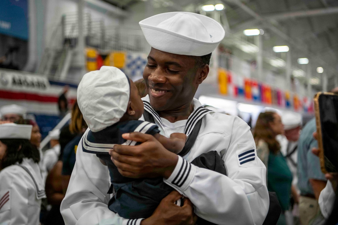 A sailor in a white uniform holds a baby wearing a sailor's uniform as fellow service members and loved ones gather in the background.