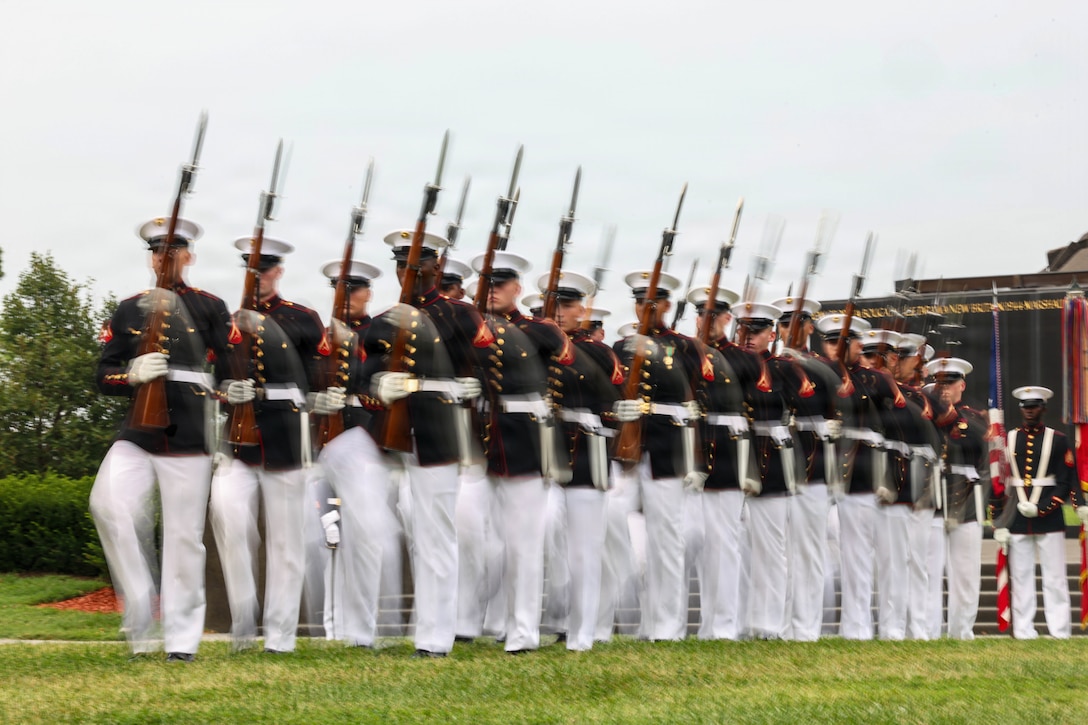 A blurred image of Marines in ceremonial uniforms marching with rifles in a line on a field during the day.