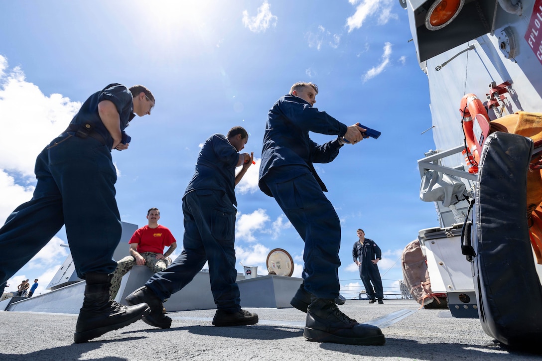 Three sailors aim weapons while moving in a triangular formation aboard a ship as fellow sailors watch during the day.