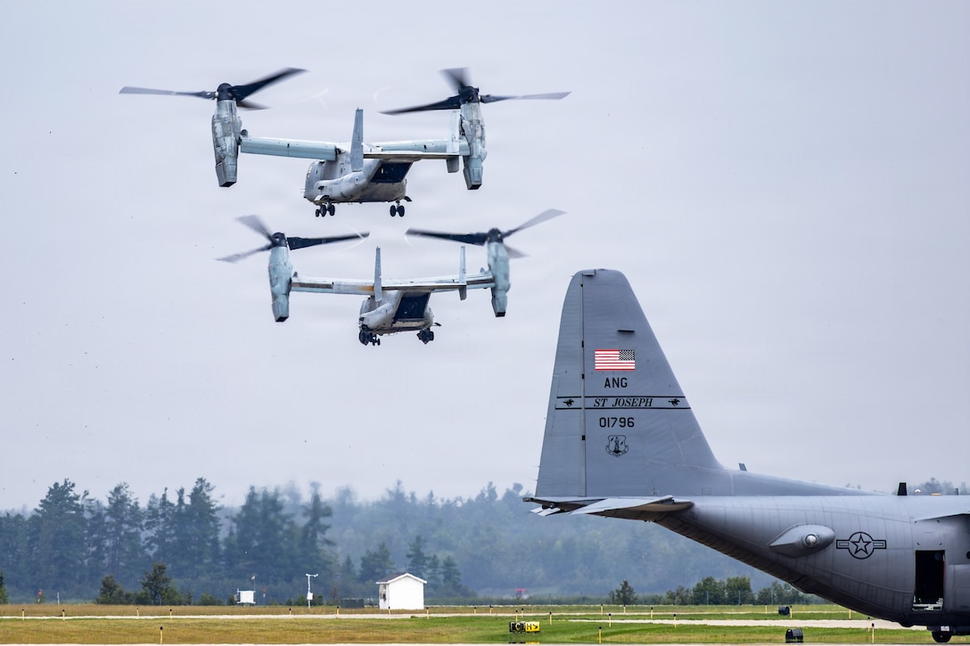 Two small aircraft with rotors at the end of each wing fly above a runway. Another aircraft is on the ground.