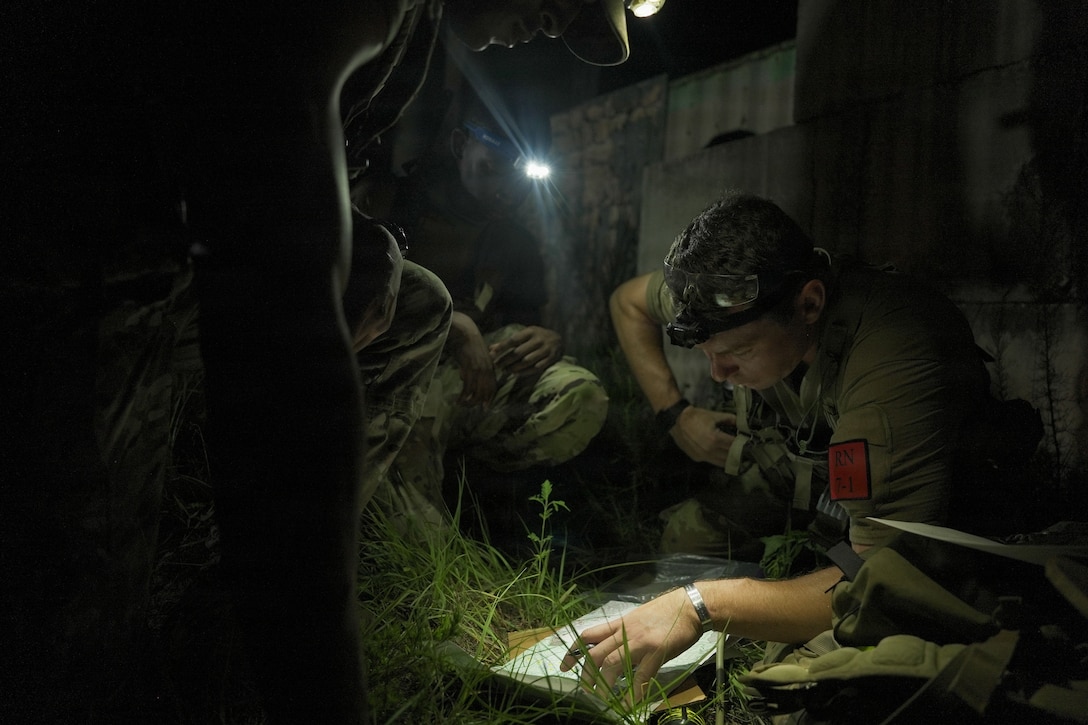 A soldier reads a paper while sitting in the grass at night. Other soldiers shine headlamps on the paper.
