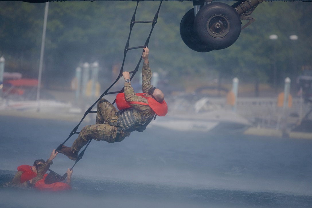 People in camouflage uniforms and red puffy vests climb out of water onto a flexible ladder connected to an aircraft.