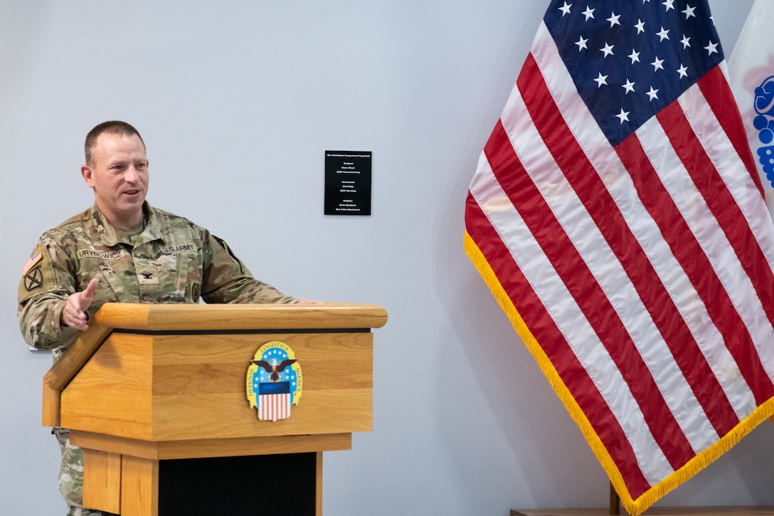 A man wearing a camouflage military uniform stands behind a podium speaking.