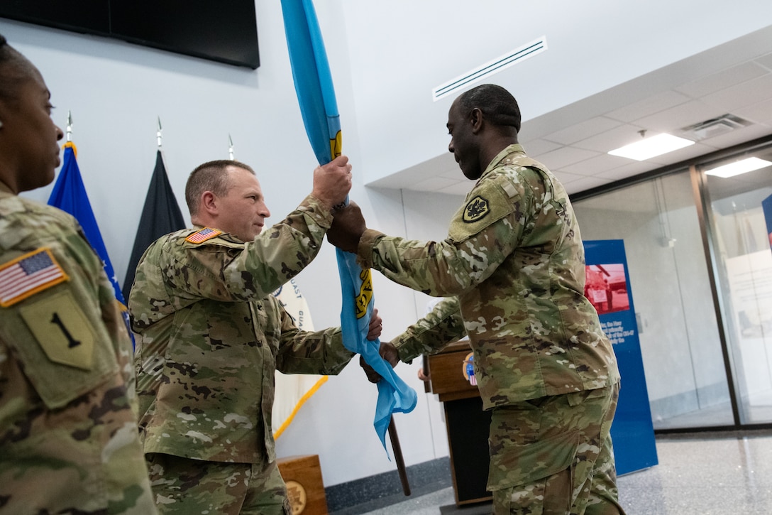 two men wearing camouflage military uniforms pass a flag to each other