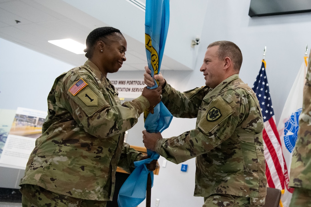 A woman wearing a camouflage military uniform takes a flag from a man wearing a camouflage military uniform.