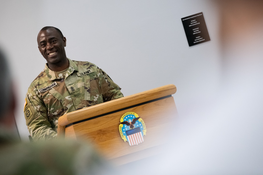 A man wearing a camouflage military uniform stands behind a podium speaking.