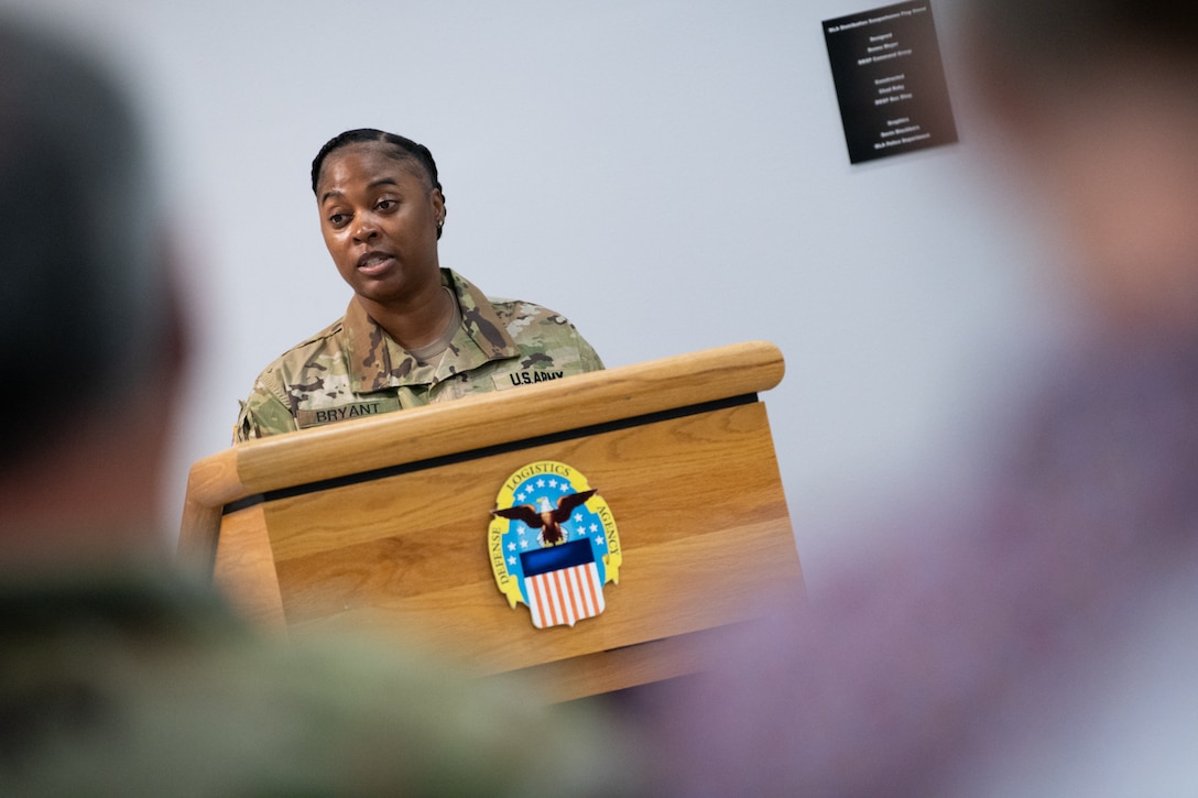 A woman wearing a camouflage military uniform stands behind a podium speaking.
