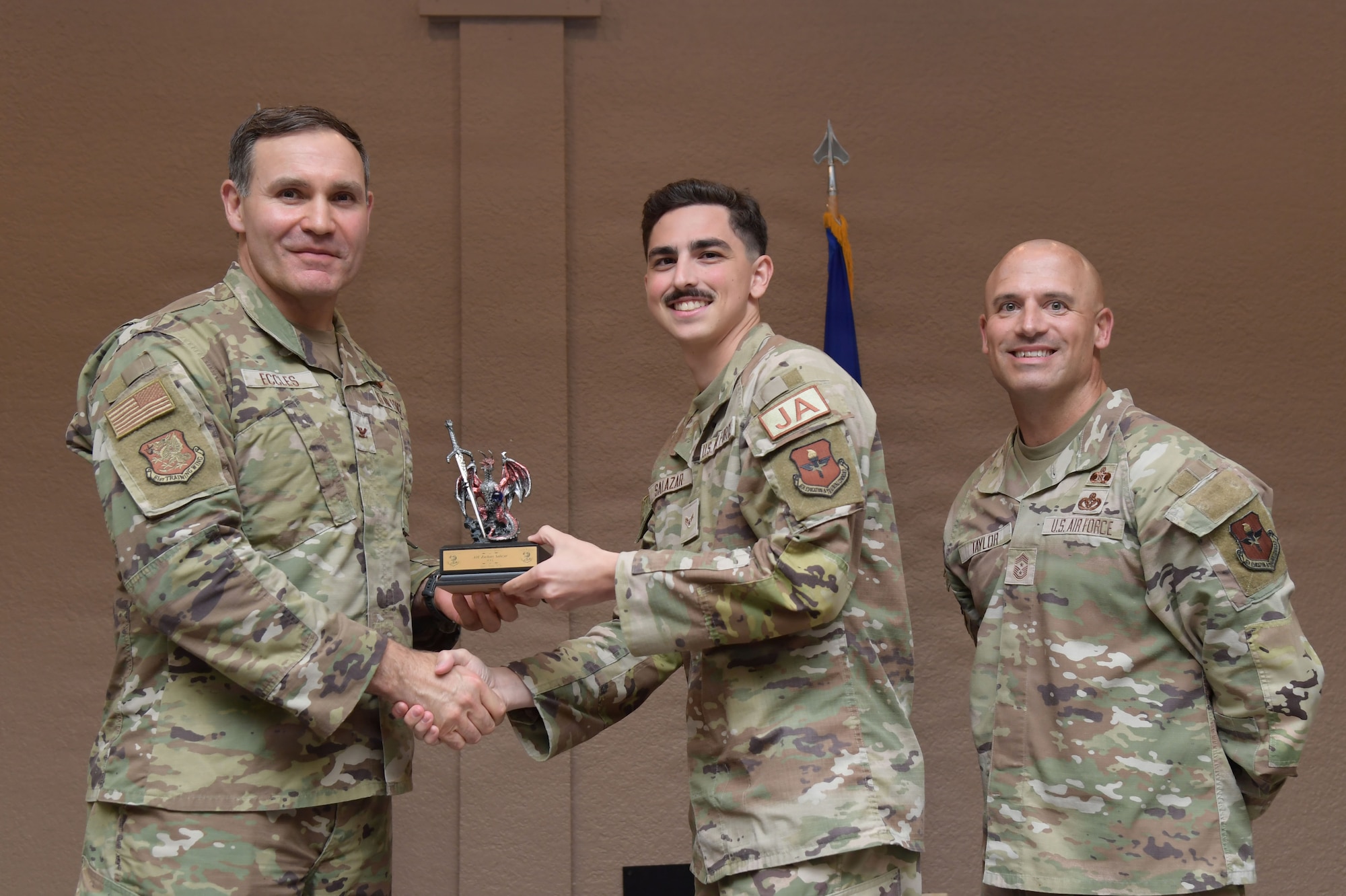 Three men wearing U.S. Air Force uniforms stand on stage holding a certificate.