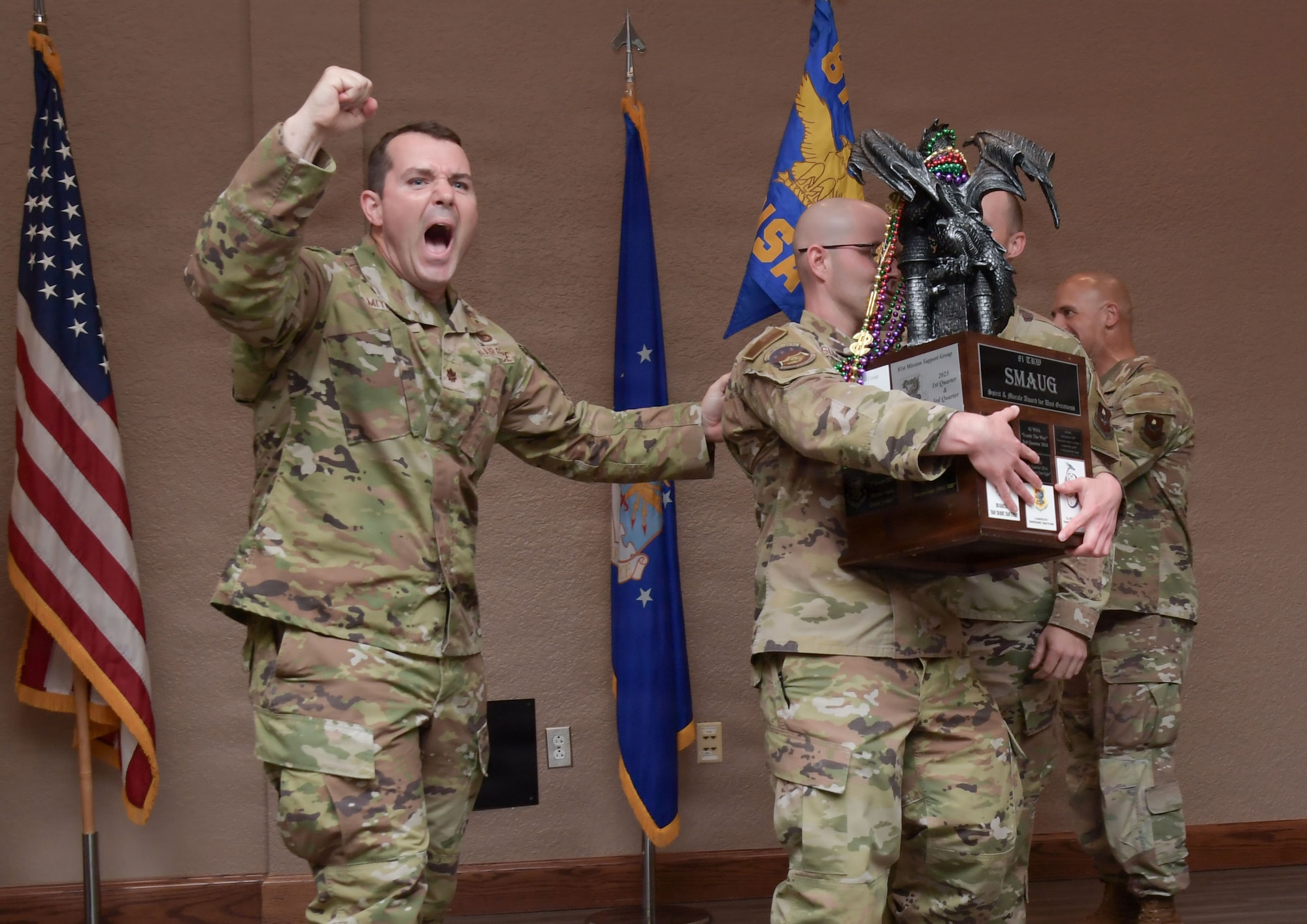 A man wearing a U.S. Air Force uniform cheers while another man holds a large trophy.