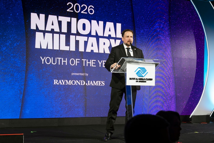 A man in business attire stands at a lectern and speaks into a microphone. On the wall behind him are the words "2026 National Military Youth of the Year."