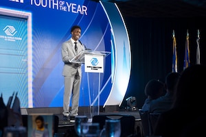 A young man in business attire stands at a lectern and speaks into a microphone. Behind him is a blue wall with the logo for Boys & Girls Clubs of America.