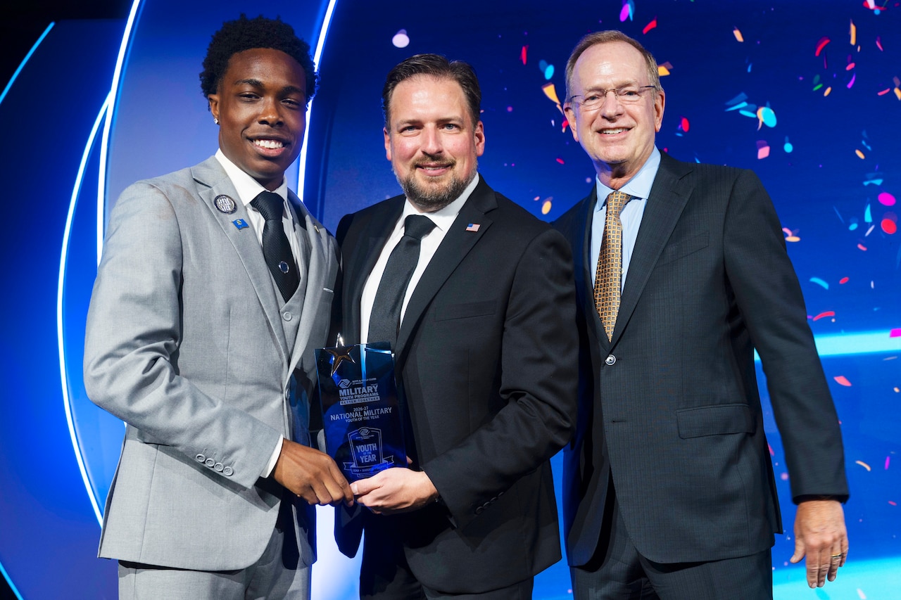 Three people in business attire stand together on a stage holding a glass award.