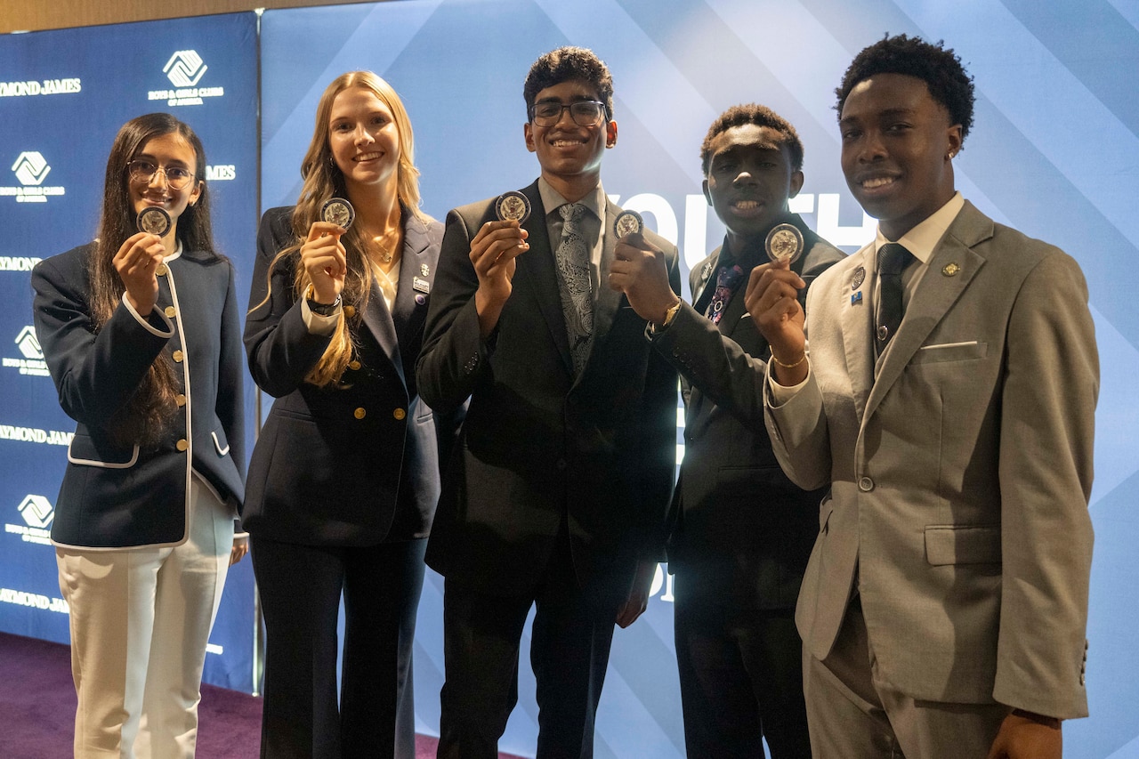 Five young people in business attire stand together and hold up commemorative coins. Behind them, a backdrop displays the logo of Boys & Girls Clubs of America.