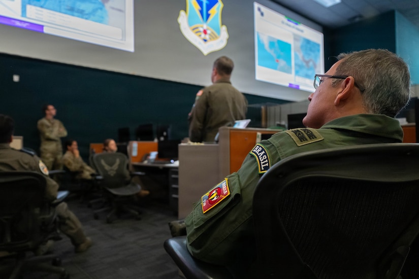 A shoulder view of a man in a military uniform sitting in an auditorium and looking up at a screen. His sleeve has various patches; one says, &ldquo;Brasil.&rdquo;