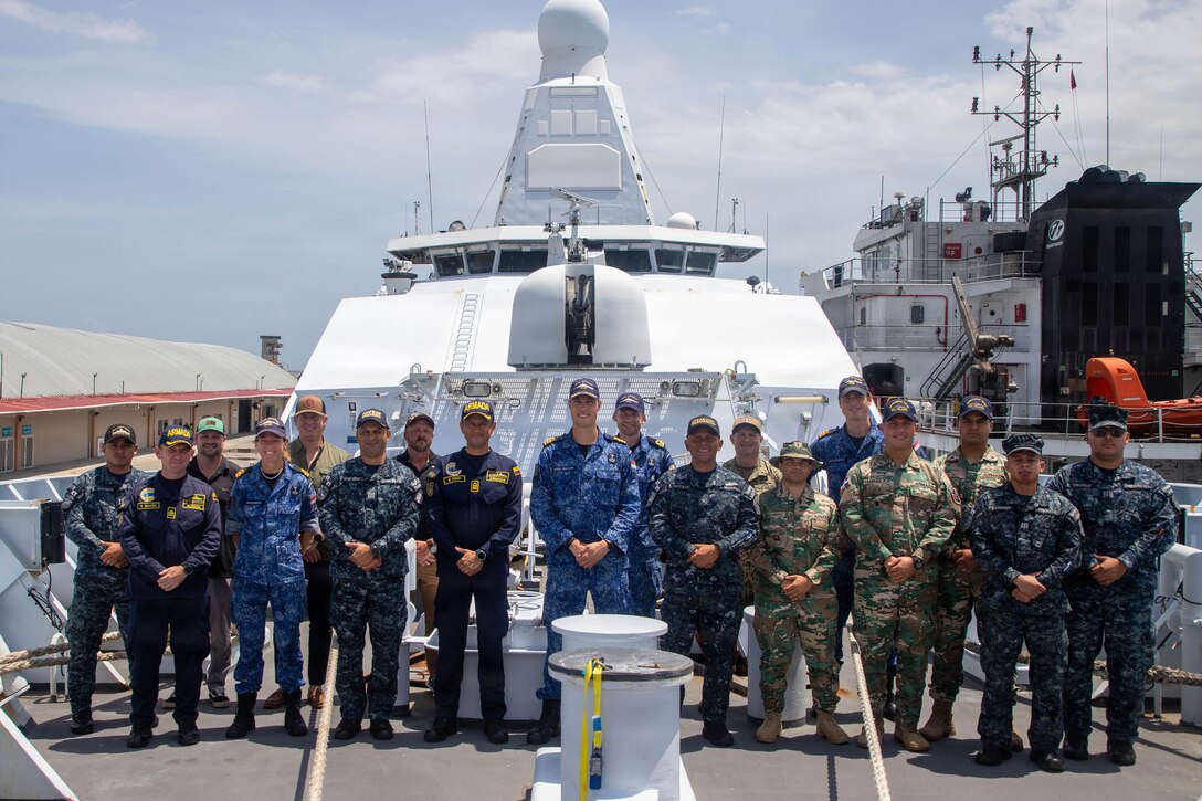 Service members in different uniforms pose for a photo on the deck of a ship at a port.