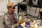 A man in casual attire and wearing eye protection sits at a table and examines a mechanical device; the table is filled with electronic equipment and lights.