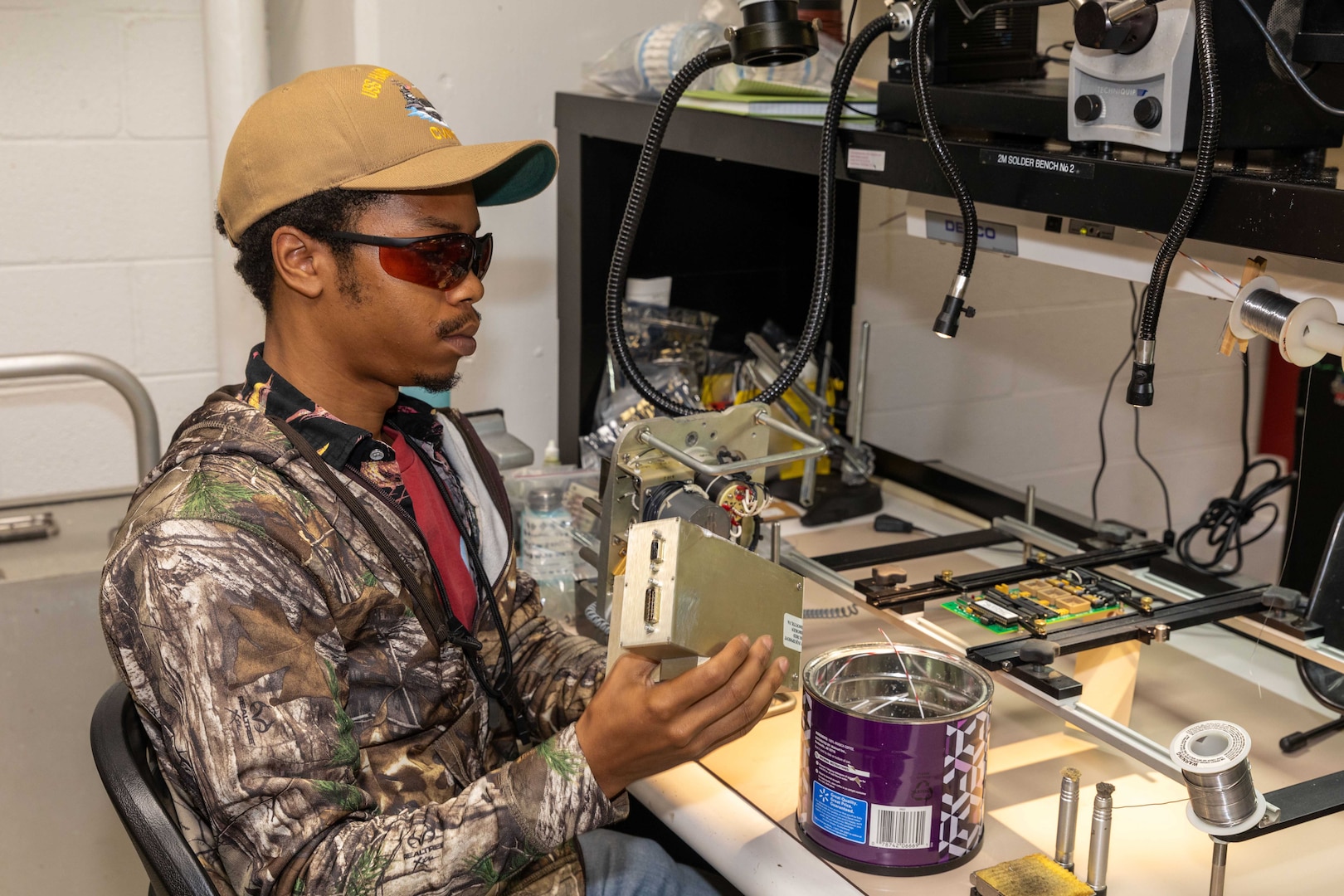 A man in casual attire and wearing eye protection sits at a table and examines a mechanical device; the table is filled with electronic equipment and lights.