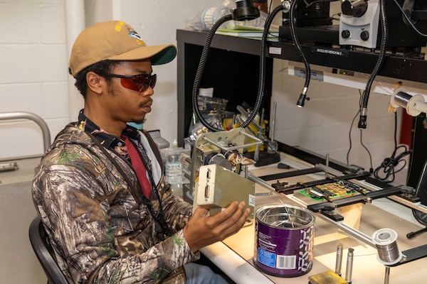 A man in casual attire and wearing eye protection sits at a table and examines a mechanical device; the table is filled with electronic equipment and lights.