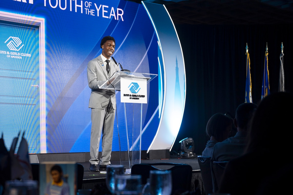 A young man in business attire stands at a lectern and speaks into a microphone. Behind him is a blue wall with the logo for Boys & Girls Clubs of America.