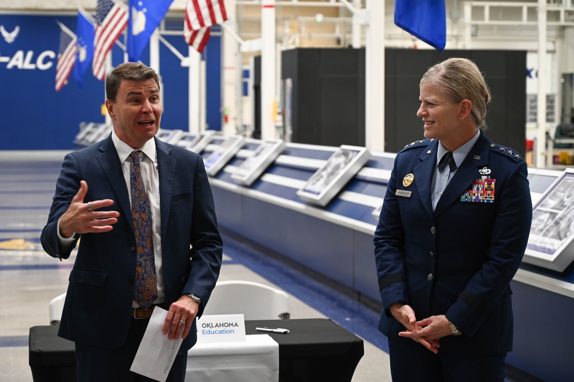 A man in a suit explains something while a person in a military uniform smiles and listens.