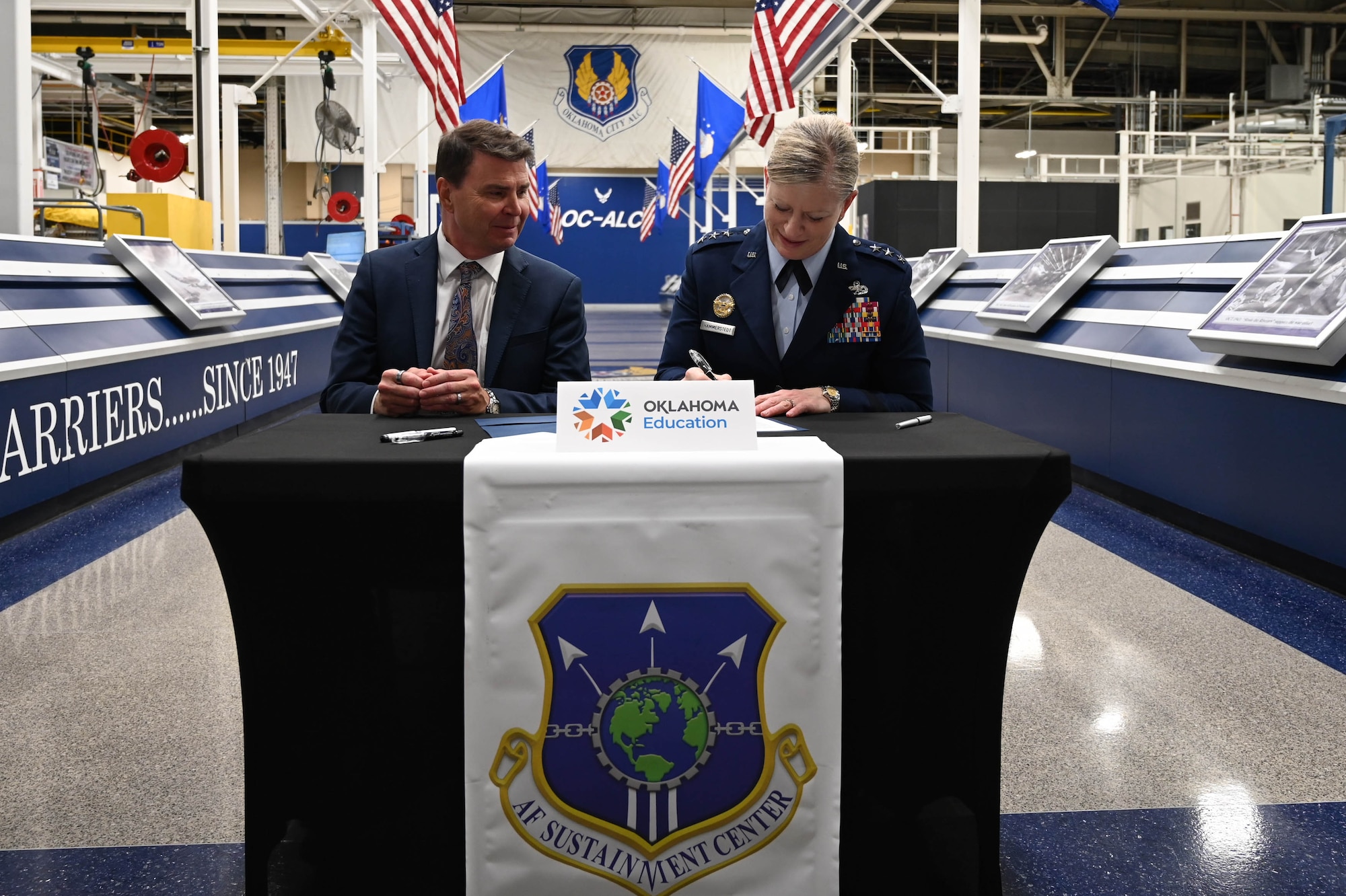 A man in a suit explains something while a person in a military uniform smiles and listens.