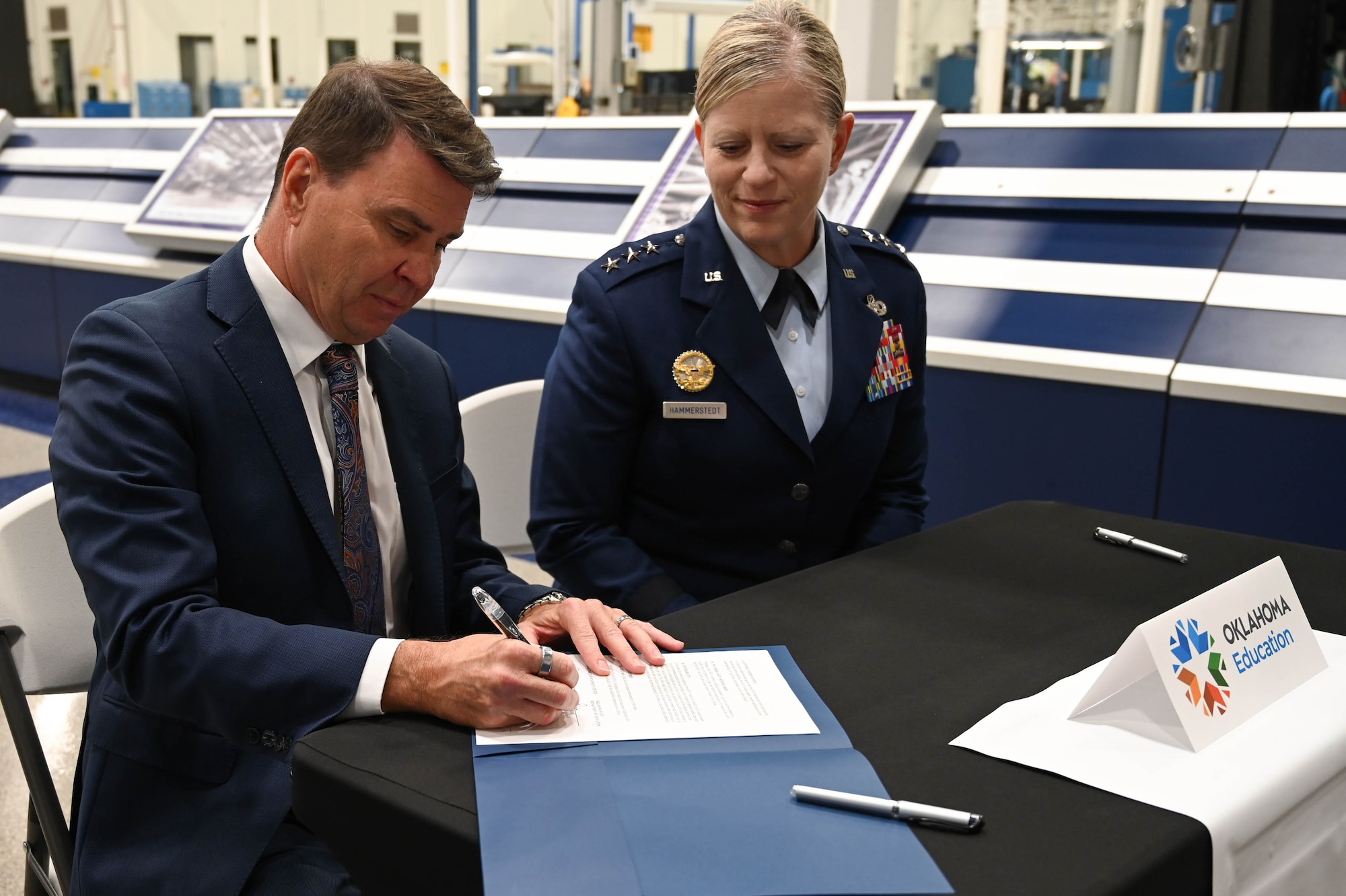 A man in a suit explains something while a person in a military uniform smiles and listens.