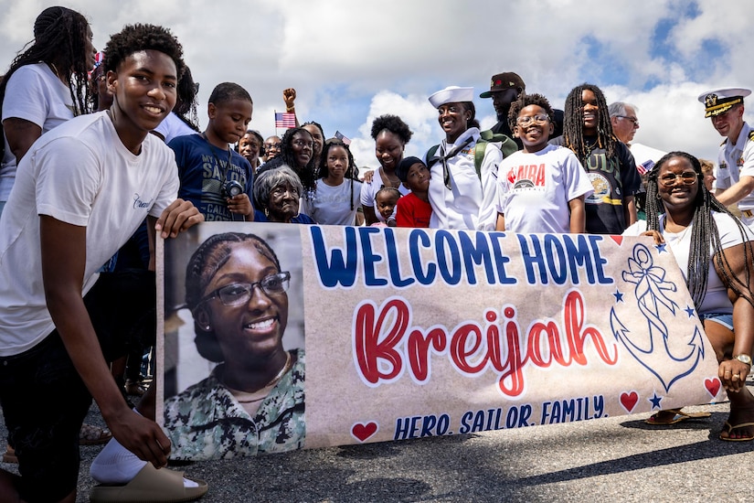 A woman in a white military uniform stands with more than a dozen people outside behind a banner that reads, "Welcome home Breijah, hero, sailor, family."