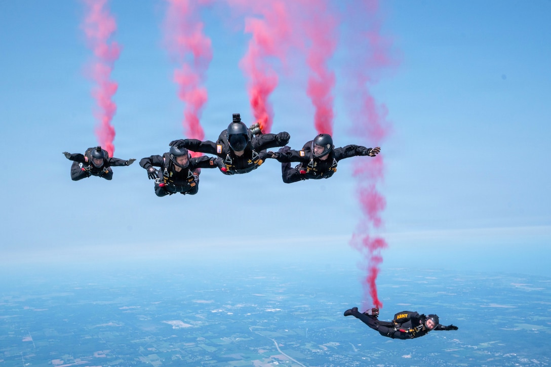 Five soldiers, four in the center and one in the foreground, descend over a city's landscape while leaving trails of red smoke in a blue sky.