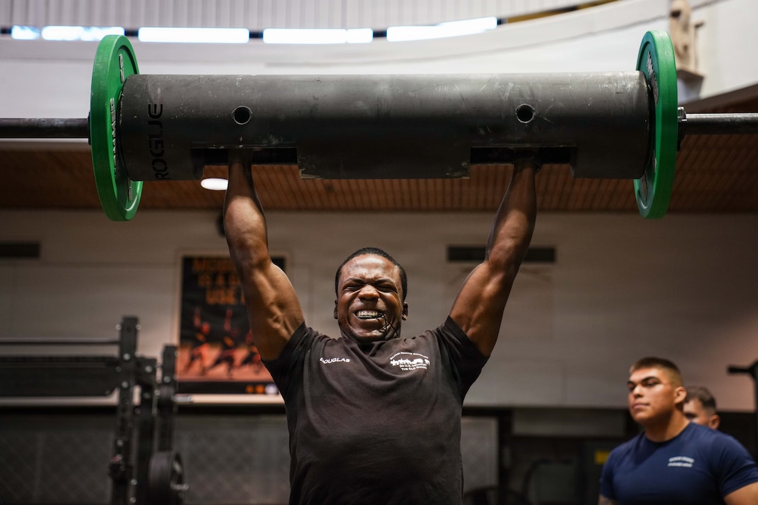 A soldier strains while lifting heavy weights into the air in a dimly lit gym as fellow soldiers watch from the side.