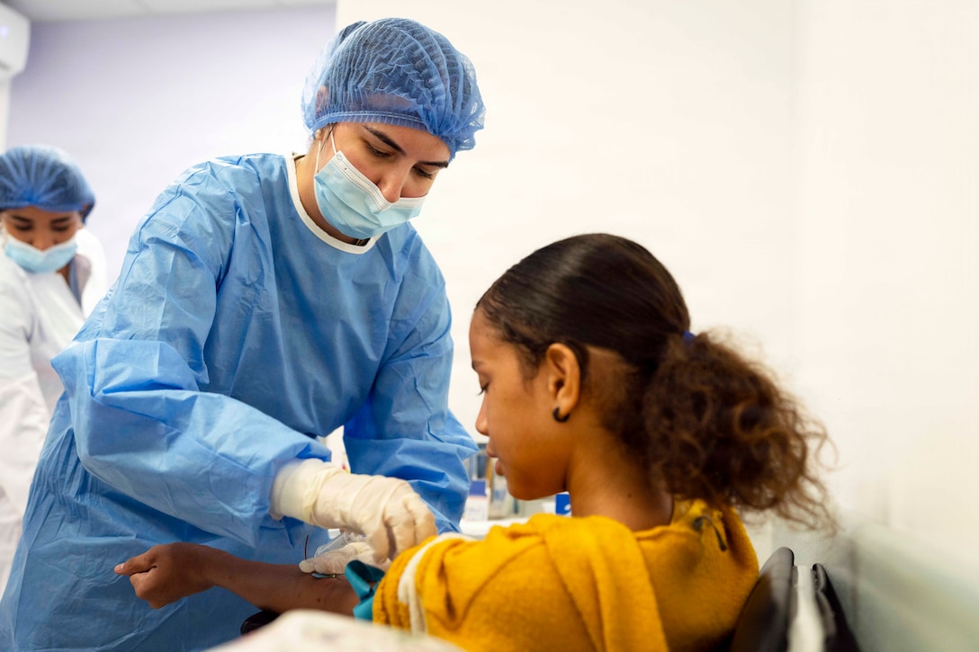 An airman in blue scrubs and a face mask takes blood from a seated patient.