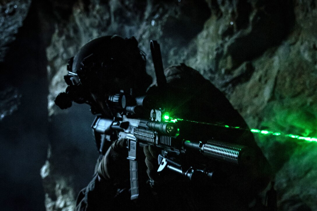 A green laser beam shoots out from the sight on a rifle held by a service member in a dark environment within a cave.