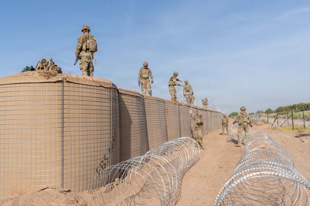Three soldiers standing on the ground hand coiled wire to soldiers standing on top of a barrier wall.