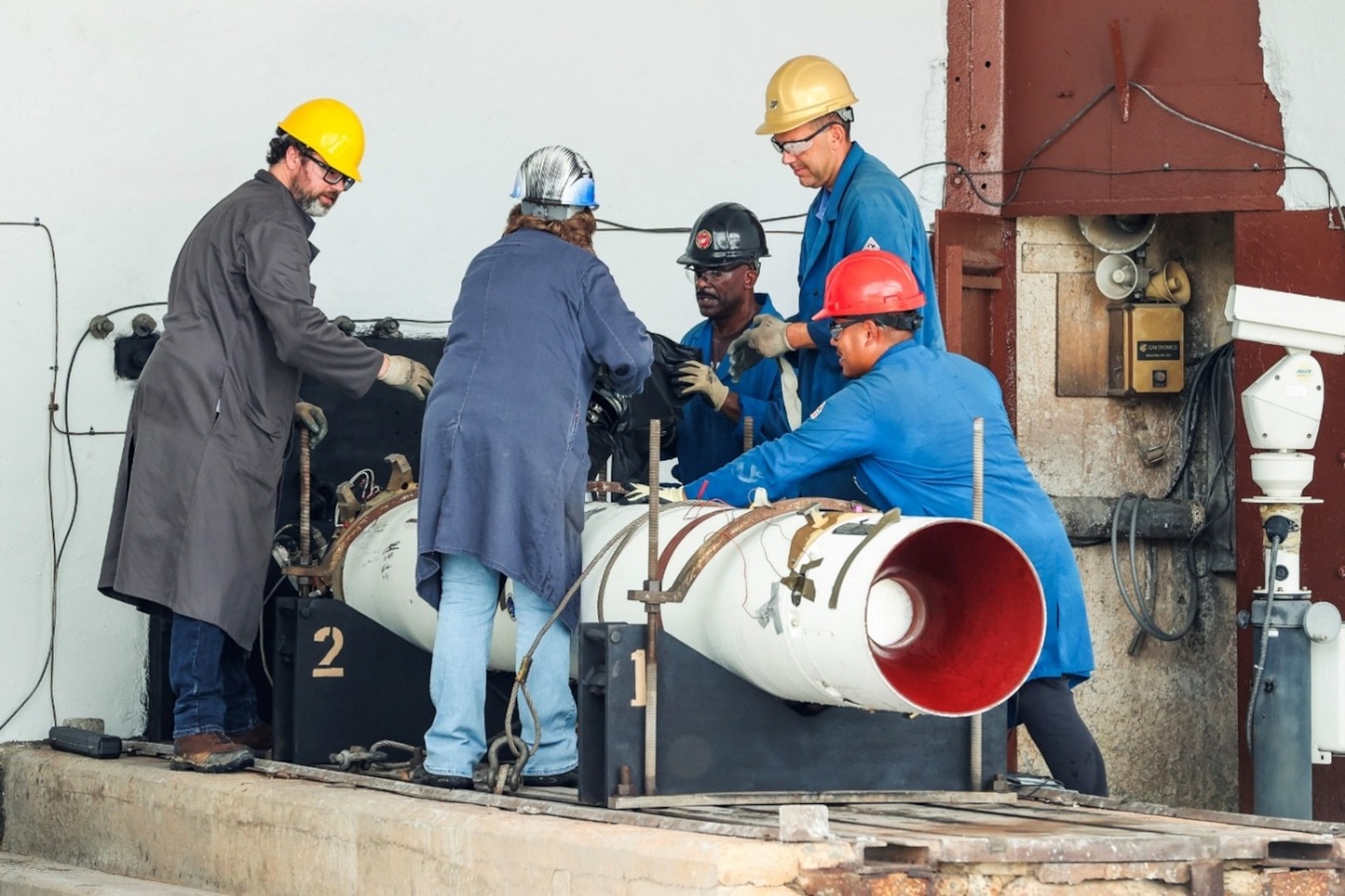 Photograph showing five workers wearing safety helmets and protective clothing collaborating around a large white cylindrical pipe with a red interior, likely part of an industrial or engineering project. Workers appear focused on assembling or inspecting the pipe, with visible equipment and structural elements in an indoor facility setting.