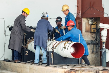 Photograph showing five workers wearing safety helmets and protective clothing collaborating around a large white cylindrical pipe with a red interior, likely part of an industrial or engineering project. Workers appear focused on assembling or inspecting the pipe, with visible equipment and structural elements in an indoor facility setting.
