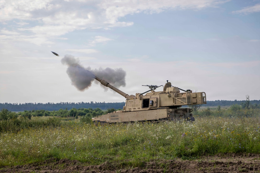 A projectile shoots out of a howitzer in a field. Trees and a cloudy blue sky are in the background.