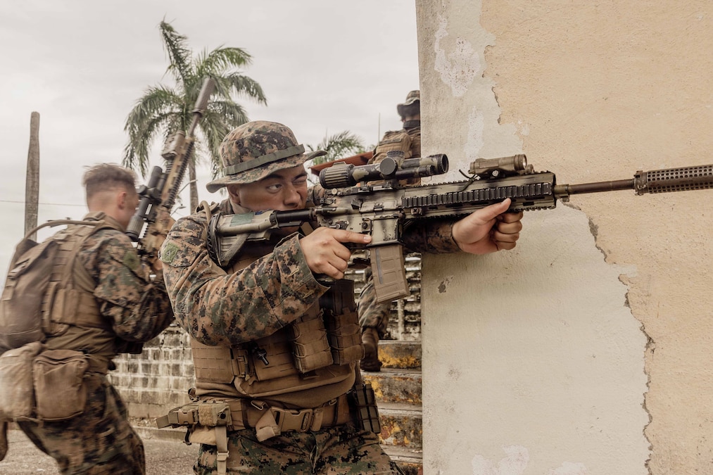 A service member in camouflage points a rifle straight ahead by the side of a wall. Two other service members facing right and a palm tree are in the background.