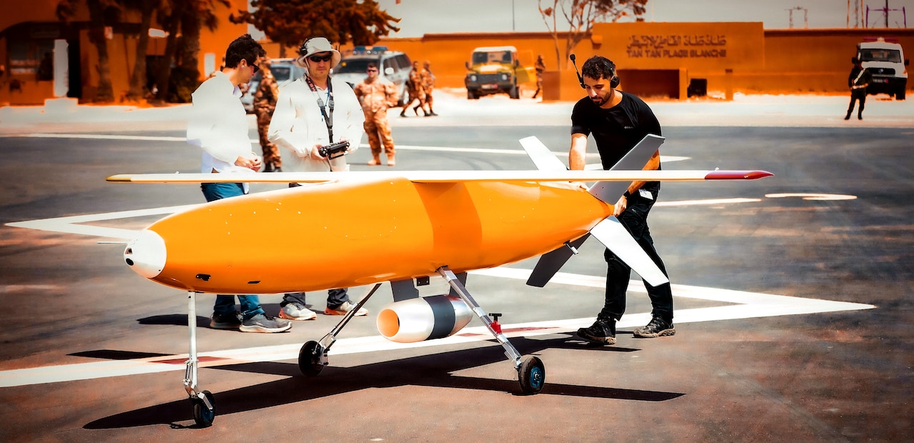 A man in casual attire pushes a large yellow and white missile in a parking lot as two other people observe.