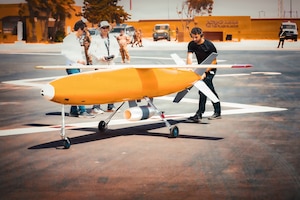 A man in casual attire pushes a large yellow and white missile in a parking lot as two other people observe.