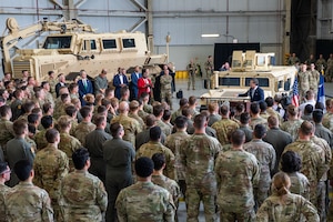 Dozens of people in camouflage military uniforms and business attire stand around a military vehicle in a hangar as a man in a suit speaks to them.