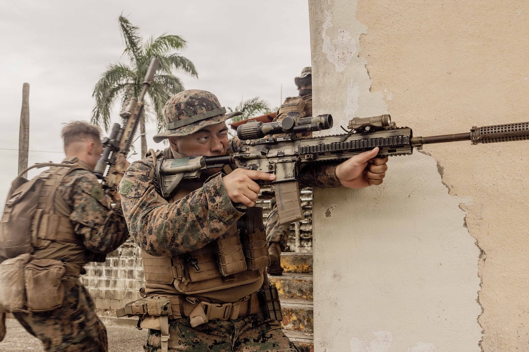 A service member in camouflage points a rifle straight ahead by the side of a wall. Two other service members facing right and a palm tree are in the background.