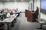 A woman speaks from behind a lectern to an audience seated in a classroom setting.
