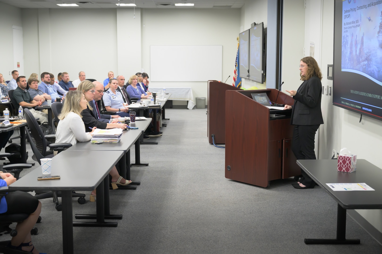 A woman speaks from behind a lectern to an audience seated in a classroom setting.