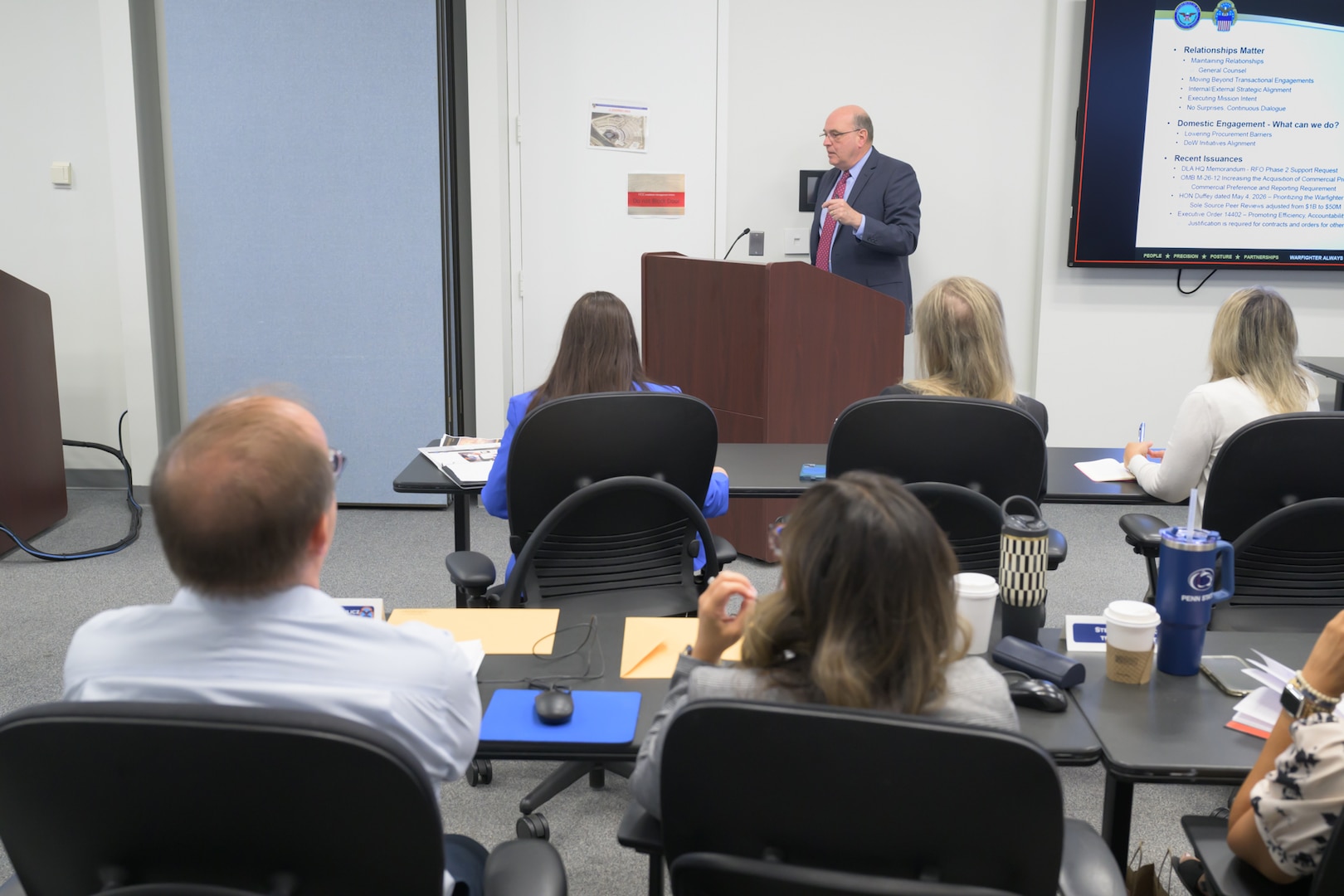 A man speaks from behind a lectern to an audience seated in a classroom setting.