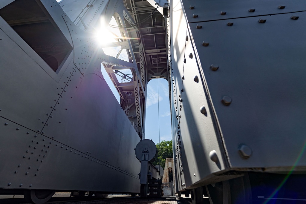 An upward-looking shot from between two massive, grey metal walls of the dam machinery, with the sun flaring brightly from behind the structure.