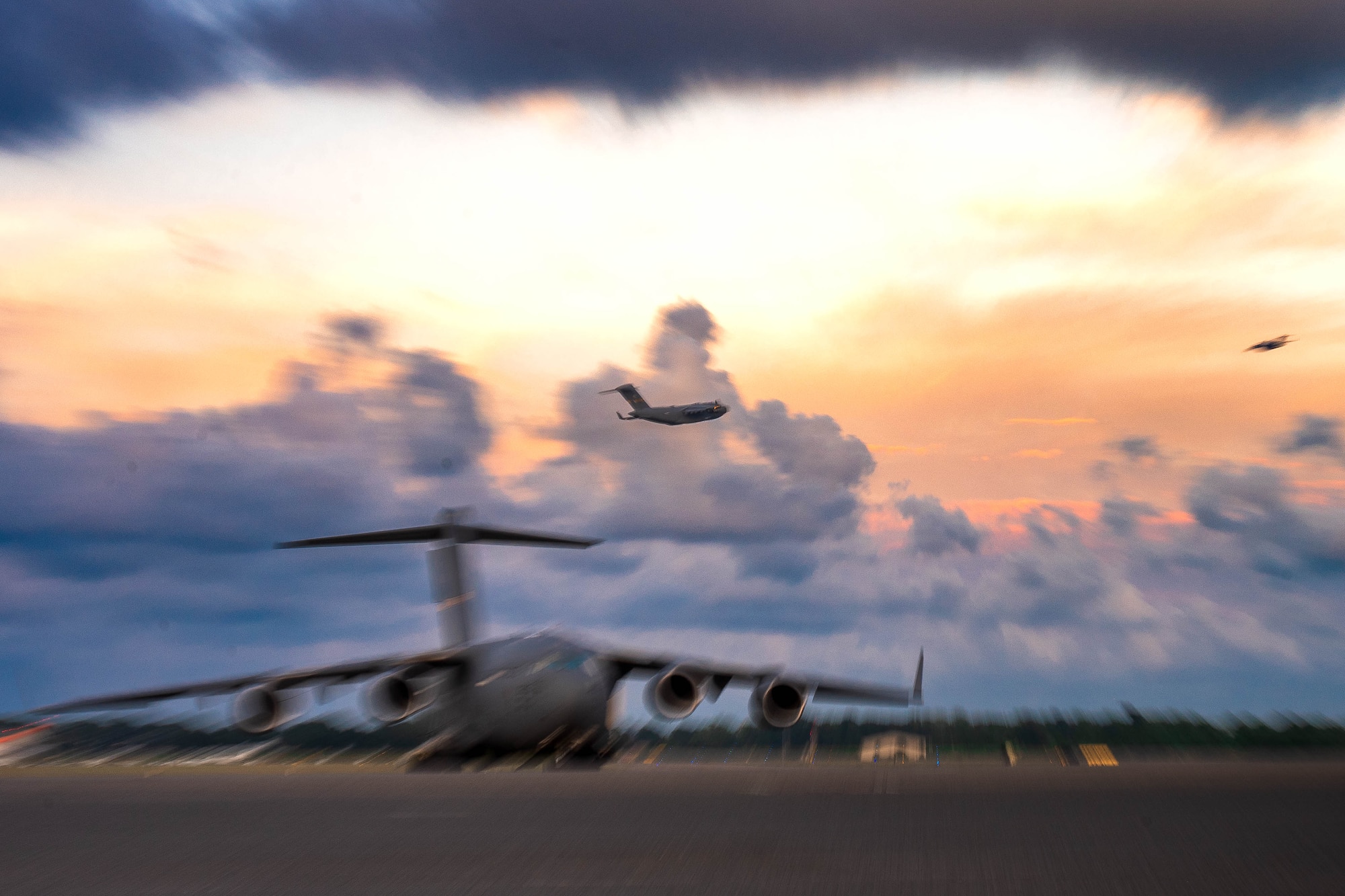 A photo of a C-17 Globemaster III flying across the sky. There's a stationary C-17 sitting on the flightline in the foreground.