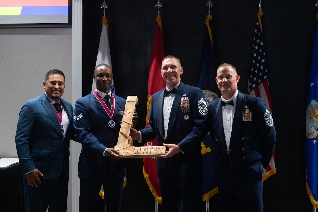 U.S. Air Force Chief Master Sgt. Derek Neill, 17th Training Wing command chief, and Senior Master Sgt. Darryl Parker, 17th Communications Squadron operations superintendent, present Airmen Leadership School Class 26-E with the “L” during an ALS graduation at Goodfellow Air Force Base, Texas, Aug. 6, 2026. Every ALS class plays a volleyball game with senior leadership and the winning team presents the “L” to the losing team at graduation. (U.S. Air Force photo by Senior Airman Brian Lummus)