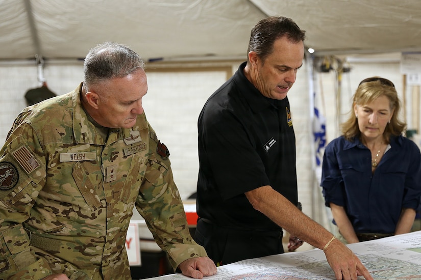 A man wearing business attire points to a map while speaking to a woman in similar attire and a man in a camouflage military uniform; they are all standing inside a large tent.