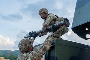 A man in camouflage military uniform bends down to take ammunition from another man in similar attire.