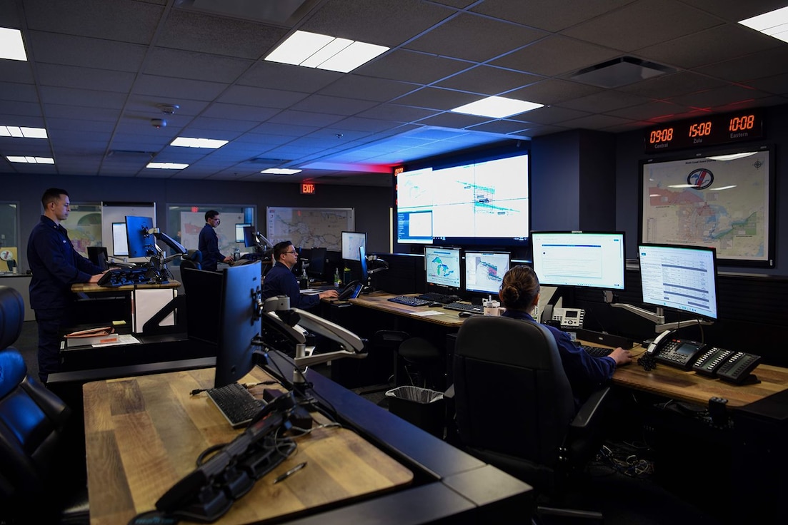 Four people standing in a control room looking at computers.