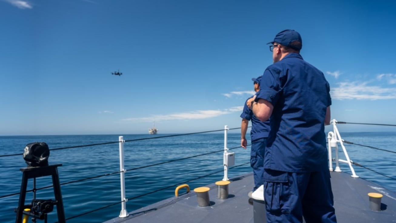 Two people watching a drone hover over the ocean.